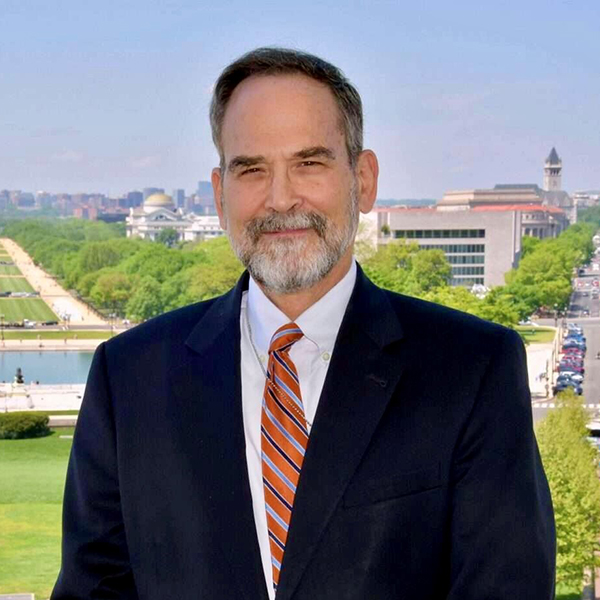 A man in a black suit, white collared shirt, and red tie, stands and smiles
