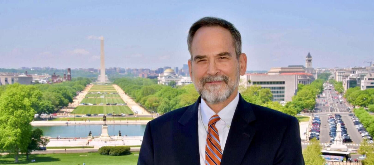 A man in a black suit, white collared shirt, and red tie, stands and smiles