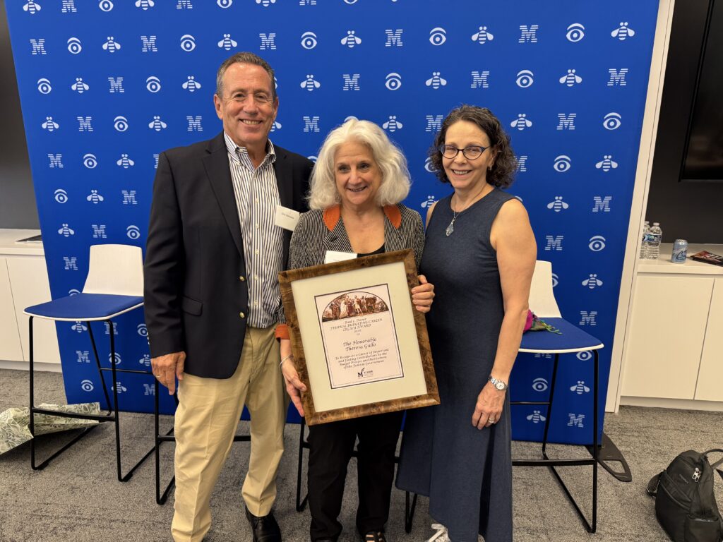Three people pose together in front of a George Mason University backdrop, with the person in the center holding a framed Paul L. Posner Federal Budgeting Career Legacy Award while the two others stand smiling beside them.